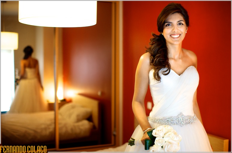 Na frente de um grande espelho e de uma parede a vermelho, a noiva a sorri, com o ramo de flores na mão e reflectida.