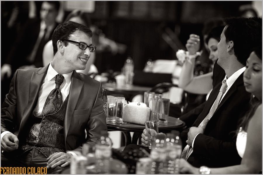 O noivo, sorrindo, sentado numa mesa conversa com convidados durante a festa do casamento.
