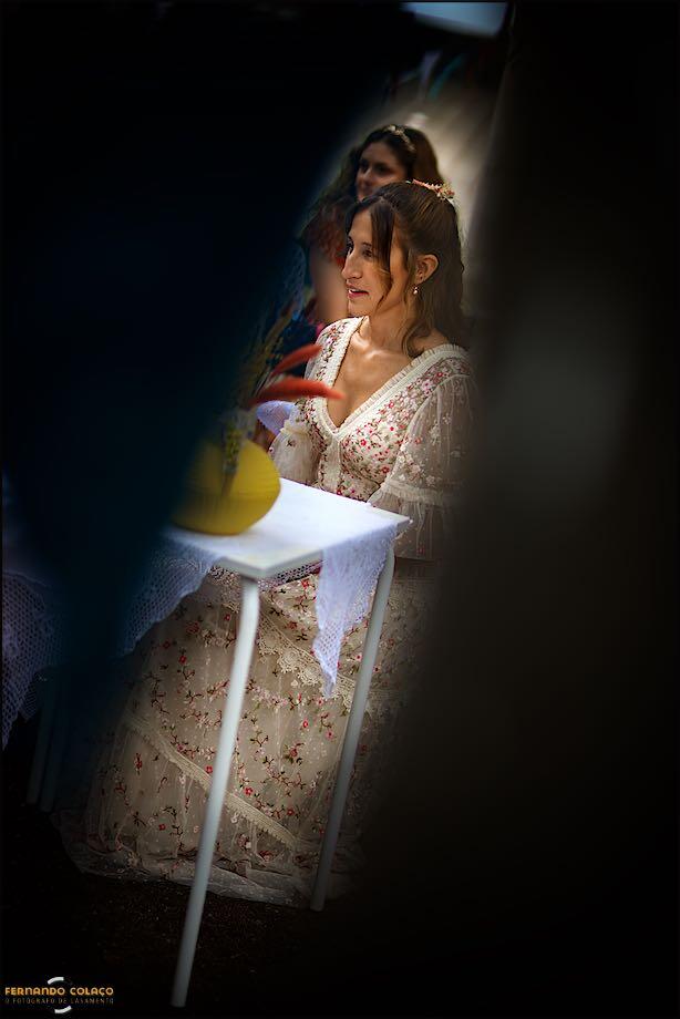 Vista por uma abertura de luz por entre dois convidados, a noiva, ainda sentada, quando acabou a cerimónia, conforme o fotógrafo de casamento em Lisboa a viu.