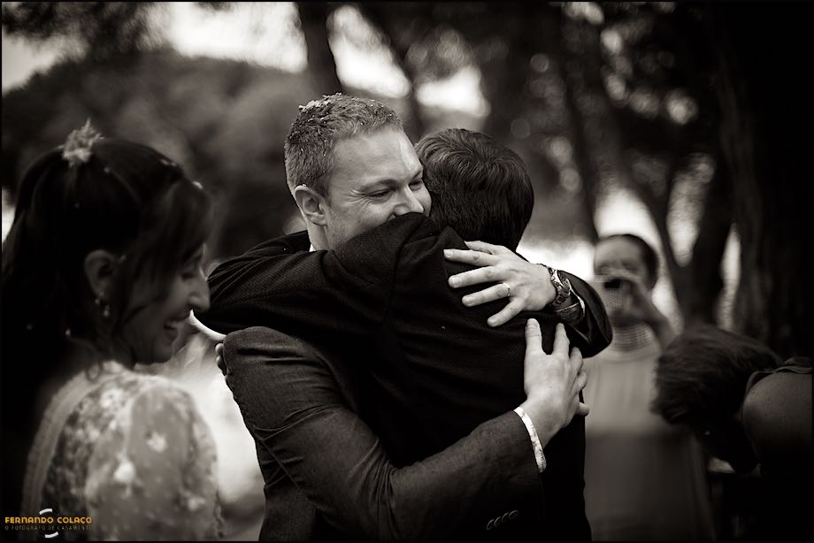 Ao lado da noiva que sorri, o noivo abraça a pai da noiva quando acabou a cerimónia do casamento, vistos pelo fotógrafo de casamento no Castelo de S. Jorge em Lisboa.