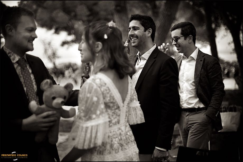 O noivo com o seu ursinho na mão em conversa com a noiva, desfocados, a serem vistos pelo padrinho do casamento numa toma do fotógrafo de casamento em Portugal.