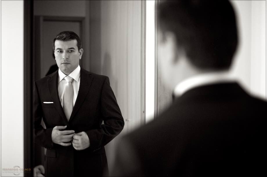 O noivo num espelho aperta os botões do casaco do fato, quase pronto para partir para a cerimónia, visto pelo fotógrafo de casamento em Braga.