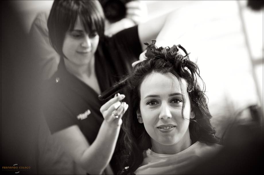 Com a cabeleireira por detrás dela, a noiva num espelho durante o penteado para a cerimónia, numa composição do fotógrafo de casamento em Braga.