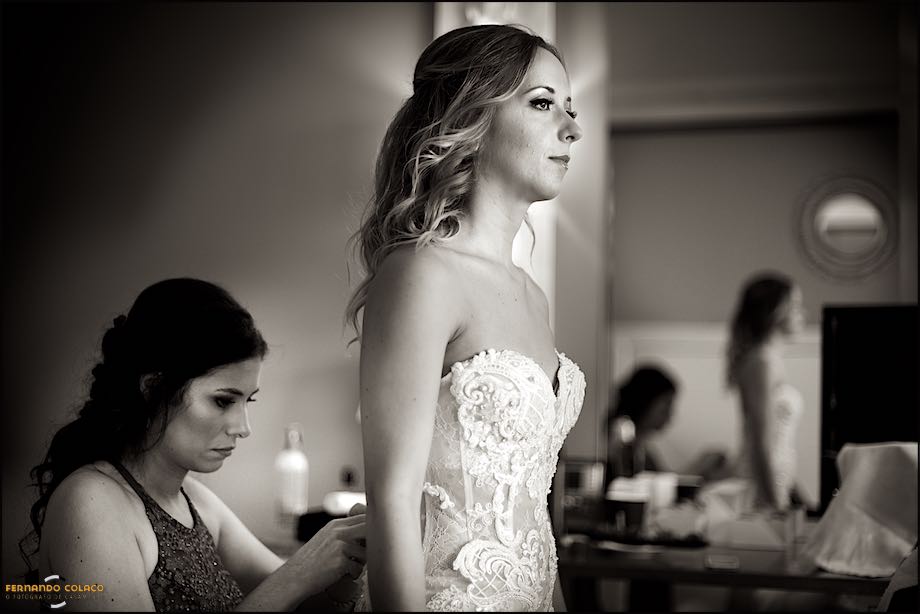A noiva quando uma amiga lhe aperta o vestido, reflectidas, mas desfocadas, num espelho, durante a preparação para a cerimónia, vistas pelo fotógrafo de casamento em Cascais.