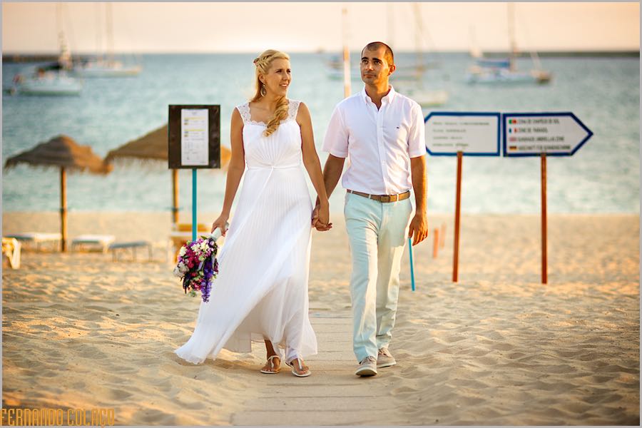 Na praia em frente ao Club Nau em Ferragudo, os noivos caminham de mãos dadas, com o mar com barcos pelas costas, vistos pelo fotógrafo de casamento no Algarve.