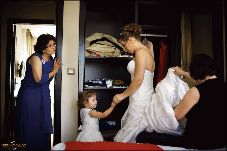 While a friend helps the bride with her wedding dress, her mother chats with her and her little daughter shows her something in her hand, all in a composition by the wedding photographer in Lisbon.