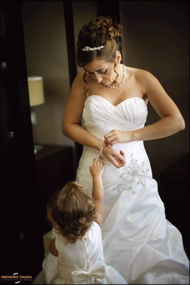 The bride clasping the bracelet on her wrist with her little daughter wanting to help her by stretching her arm, seen by the wedding photographer in Lisbon.