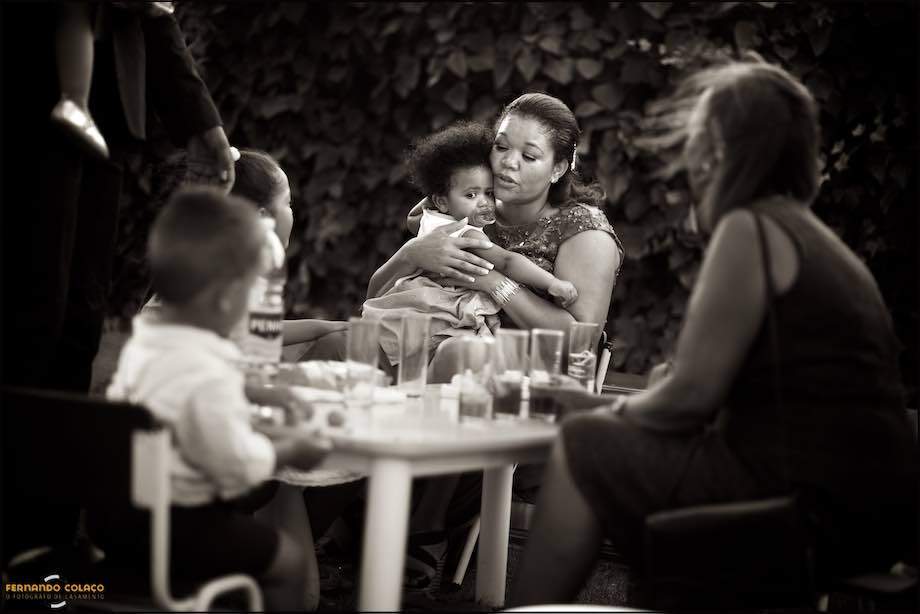 At a table in the garden where the wedding party takes place, a mother lovingly soothes her little daughter on her lap, as seen, at that moment, by the wedding photographer in Sintra.