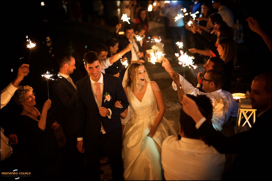 The groom and bride walk towards the wedding cake amidst golden light from glowing sticks held by guests, seen by the wedding photographer at Quinta do Castro in Cadaval.