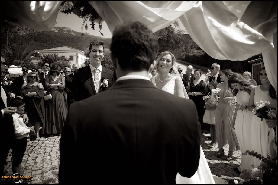 The couple at the beginning of the wedding ceremony with the officiant, from the back, and the guests behind them at Quinta do Castro in Cadaval.