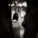 The bride with her father on their way to the wedding ceremony site, seen through the trunk of an olive tree at St. George's Castle, by the Lisbon wedding photographer.