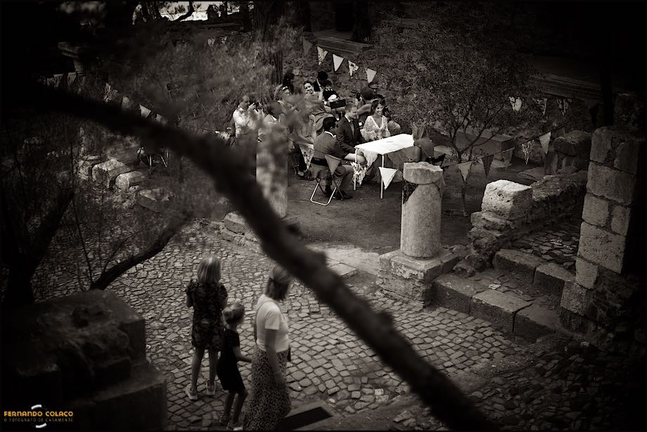 Seen from above, the bride and groom and their family members at the wedding ceremony, seen by three people passing by, at St. George's Castle in Lisbon.