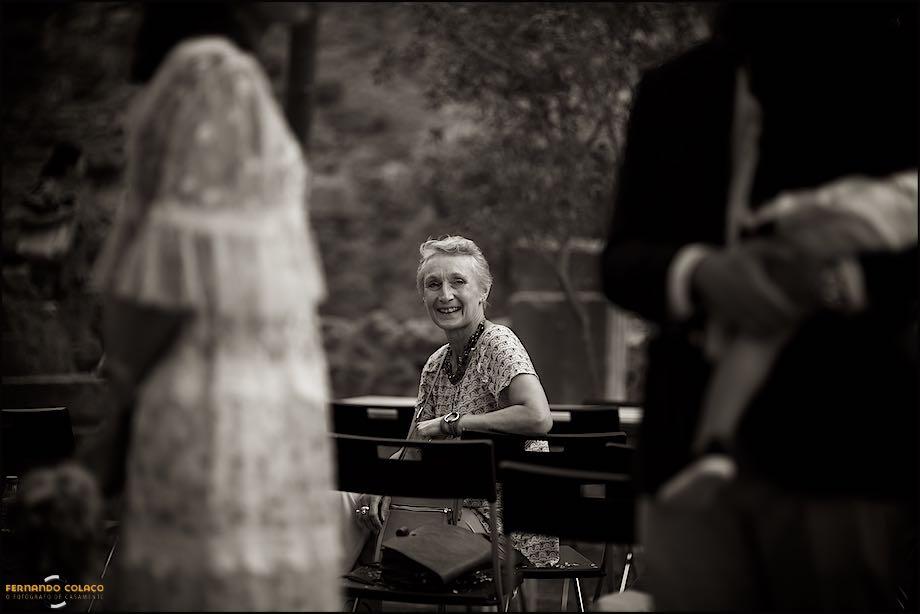 Among the blurry bride and groom, the groom's mother, sitting on a chair, laughs as she talks to them moments before they leave for the commemorative meal of the wedding, by the wedding photographer in Lisbon.