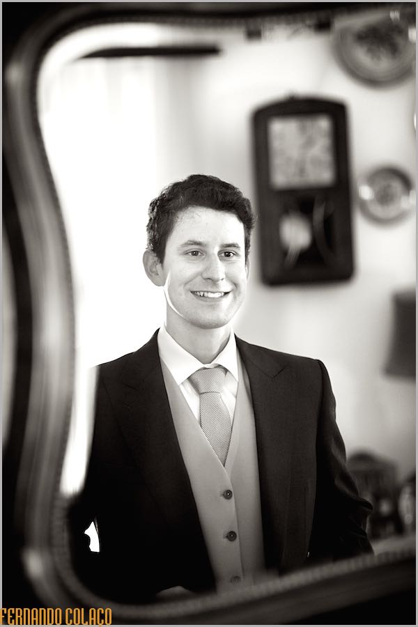 The groom smiles, already ready to leave for the ceremony, in a mirror, checking if everything is as he likes, seen by the wedding photographer in Campo Maior.
