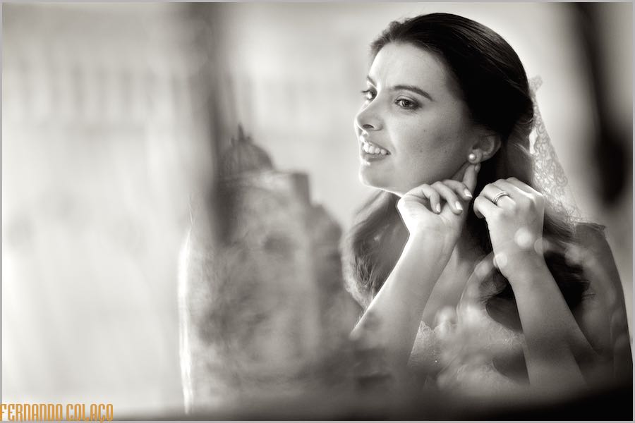 In a mirror, the bride puts one of the earrings, next to objects, blurred.