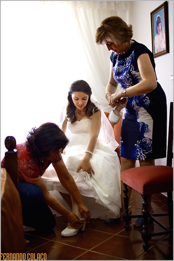 The bride sitting as her mother and an aunt help her put on her shoes.