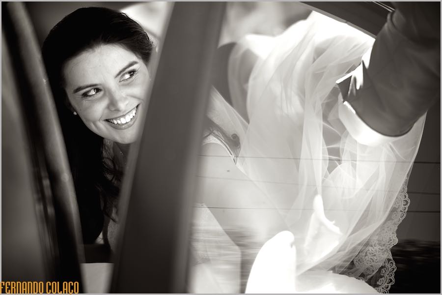 The bride smiles as she gets into the car to go to the ceremony, while a hand smoothes her dress, captured by the wedding photographer in Estremoz.