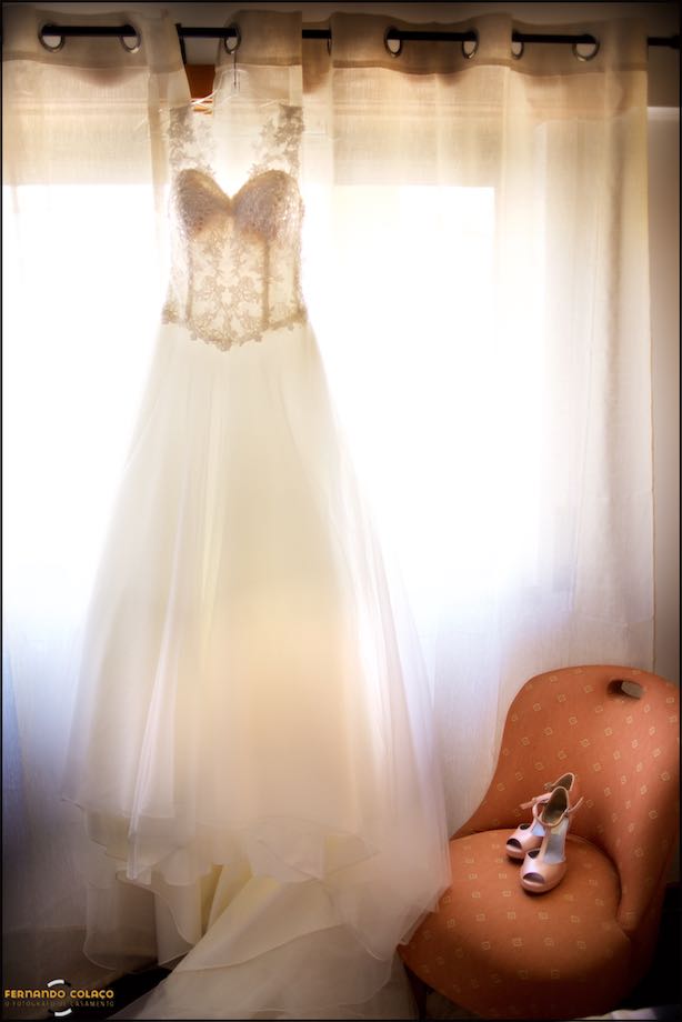 The bride's dress hanging in front of a window, next to her shoes on an armchair, in a composition by a wedding photographer in Portugal