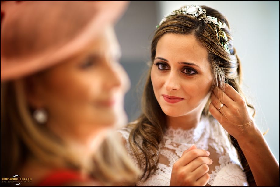 The bride with her fingers checks the positioning of an earring, next to her mother's face, in profile and blurred, as seen them by the wedding photographer in Sintra.