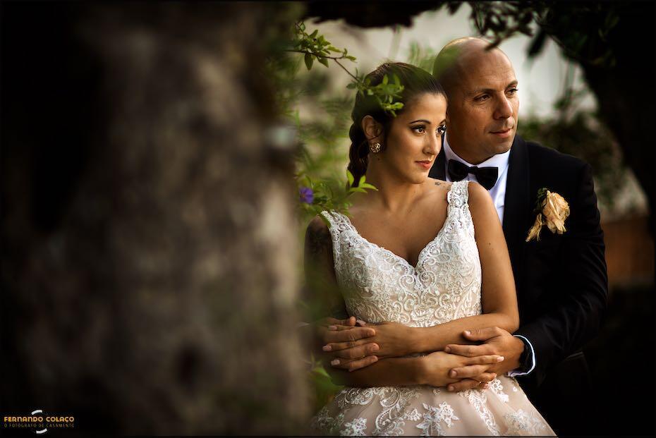 In the garden of Quinta da Ramila, the bride and groom pose, embracing, next to the trunk of an olive tree, for a photo taken by the wedding photographer in Leiria.