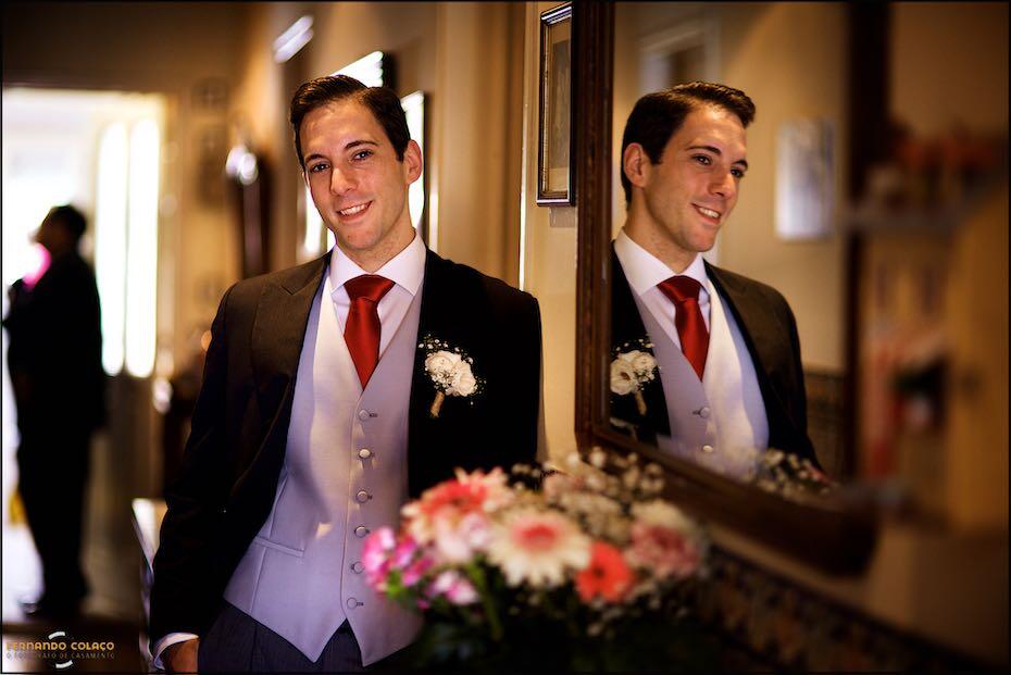 O noivo numa pose para o fotógrafo de casamento em Lisboa, na casa dos pais ao lado de um espelho que o reflecte e com um arranjo floral pela frente, desfocado.