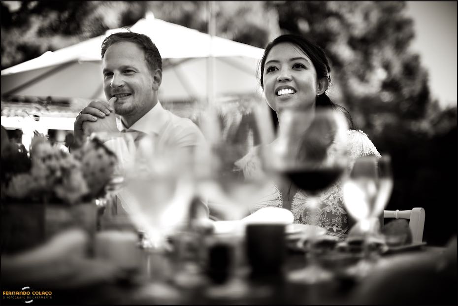 In Agostos, in the Algarve, the groom and bride listen, smiling, to a friend talking over dinner, as seen by their wedding photographer in the Algarve.