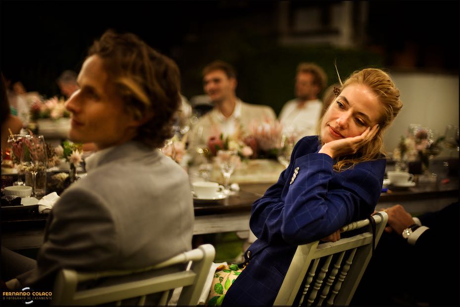 A woman wedding guest, leaning back in a chair with her hand to her face, listens intently to a speech at the wedding dinner in Agostos, as seen by the wedding photographer in the Algarve.