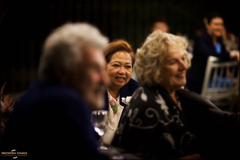 The bride's mother, seen among other blurred people, smiles as she listens to the speech at the wedding dinner in Agostos, Algarve.