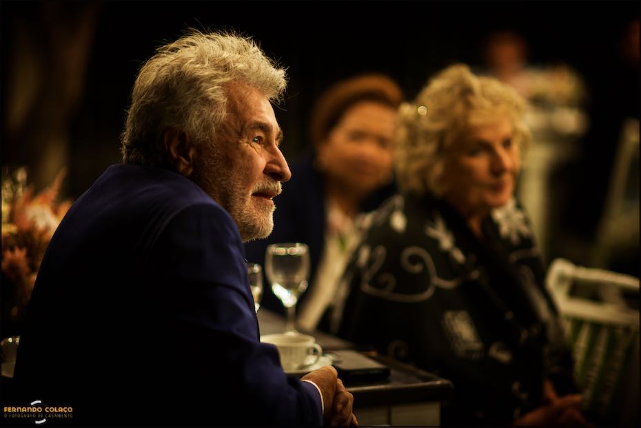 The groom's father, in profile, with his mother in the background, out of focus, listens attentively to the speaker at the dinner in Agostos, as seen by the wedding photographer in the Algarve.