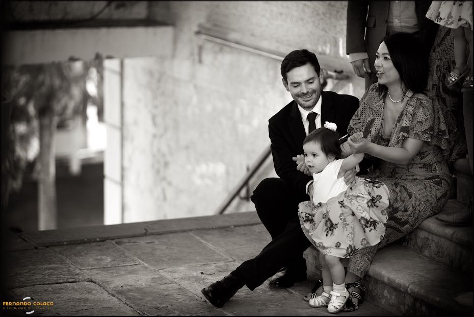 A couple, at the wedding party in Agostos, hold their baby trying to take her first steps, sitting on the steps of an old door, seen by the wedding photographer in the Algarve.