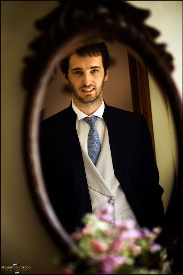 The groom looks at himself in an oval mirror with a wooden frame and a bouquet of flowers below, as seen by the wedding photographer in Évora.