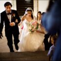 The bride and her father walk up the stairs outside the Convento do Espinheiro church, followed by the wedding photographer in Évora.