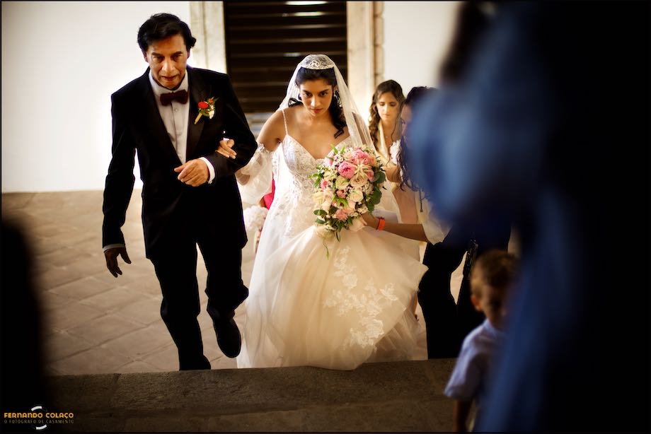 The bride and her father walk up the stairs outside the Convento do Espinheiro church, followed by the wedding photographer in Évora.