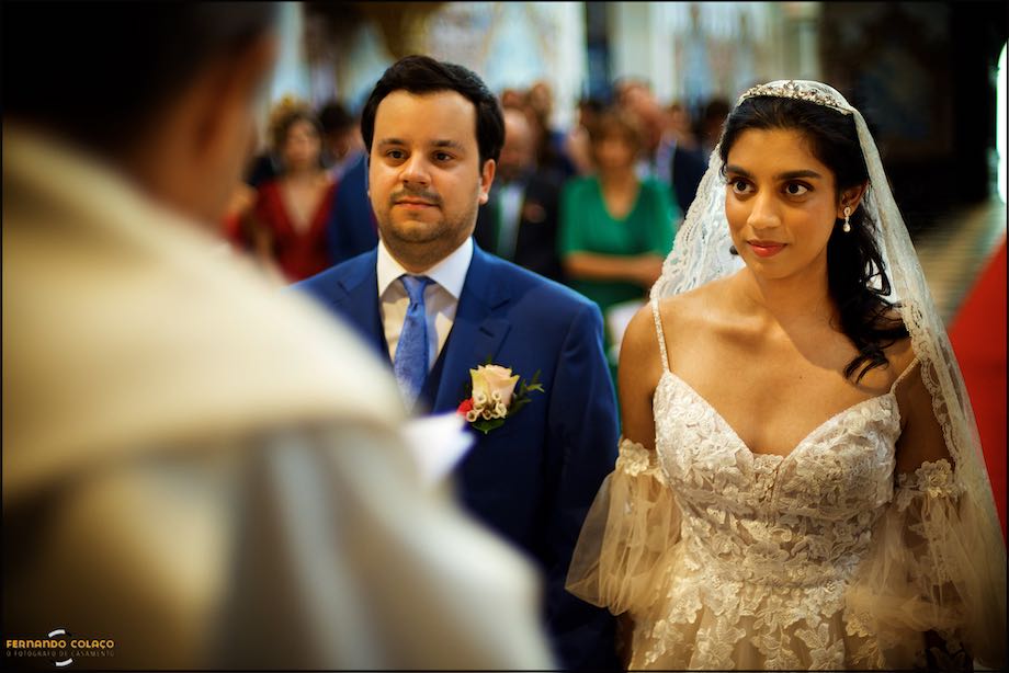 The groom and the bride, side by side, in front of the priest, out of focus and with their backs turned, during the wedding ceremony at the Convento do Espinheiro Church in Évora.