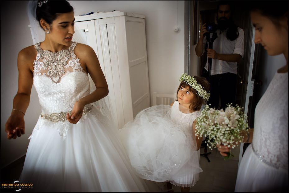 Between the bride and a friend of hers, a little girl looks very intensely at her while holding her veil, in a moment captured by the wedding photographer in Alentejo.