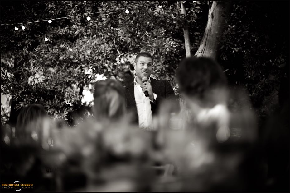 Um amigo dos noivos discursa durante o jantar do casamento nos Agostos, no Algarve, visto por entre outro convidados desfocados.