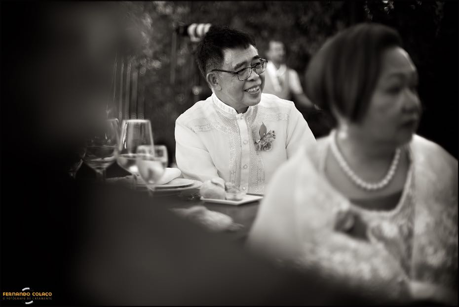 O pai da noiva, por detrás da sua mulher, desfocada, sorri enquanto, atentamente, escuta um discurso durante a festa do casamento nos Agostos, no Algarve.