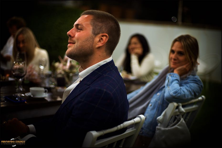 Um amigo do noivo, sentado junto de outros convidados, desfocados, sorri quando ouve um discurso no jantar do casamento nos Agostos, no Algarve.