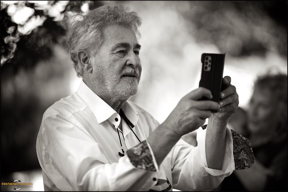 O pai do noivo no momento em que fotografa a sua mulher com a neta ao colo, na festa do casamento do filho nos Agostos, captado pelo fotógrafo de casamento no Algarve.