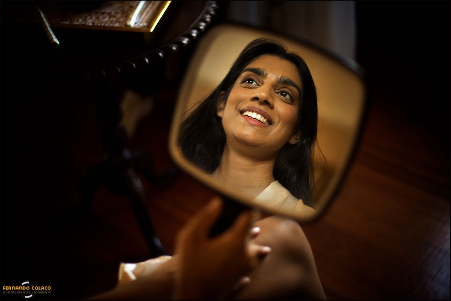 A noiva sorri, dentro de um pequeno espelho que o fotógrafo de casamento em Évora capta quando ela se está preparar para a cerimónia no Convento do Espinheiro em Évora.