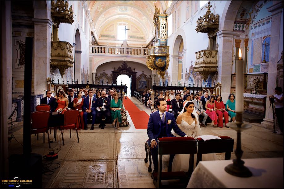Vista geral da igreja do Convento do Espinheiro em Évora, com os noivos, de frente e sentados junto do altar e todos os seus convidados por detrás, na cerimónia do casamento.