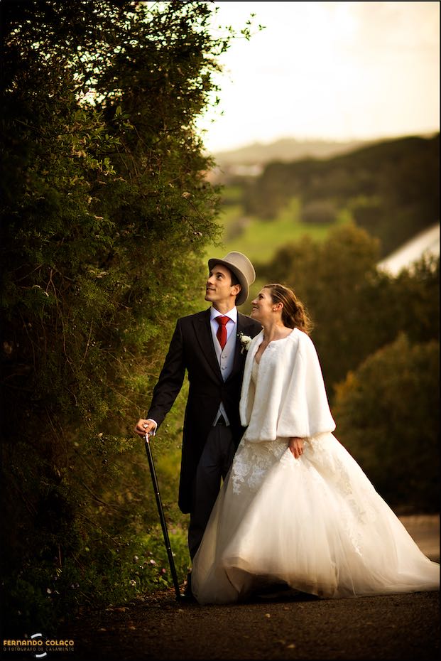 Na sessão com o fotógrafo de casamento, a noiva ao lado do noivo, de bengala e chapéu alto, junto de uma grande árvore no jardim da Casa de Reguengos.