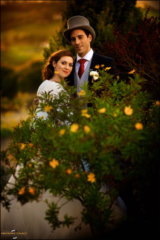Por detrás de um ramo com flores amarelas, a noiva encostada ao noivo, de chapéu alto, durante a sessão com o fotógrafo de casamento na Casa de Reguengos.