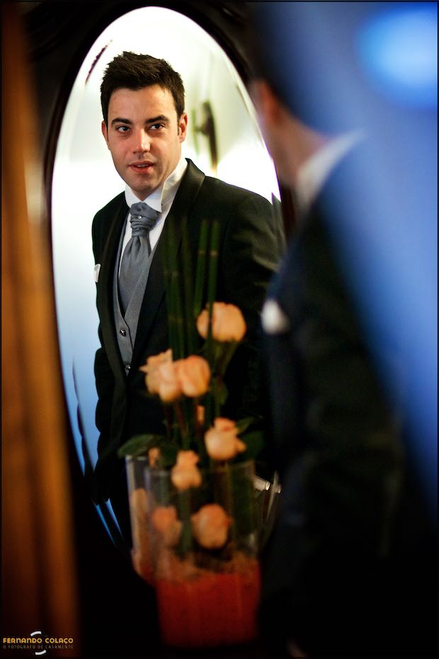 Num espelho e com uma jarra de rosas cor de laranja desfocadas, o noivo olha para si, verificando de está tudo em condições para partir para a cerimónia do casamento.