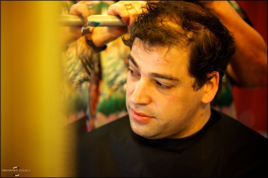 Rosto do noivo, rodeado de elementos amarelos desfocado e com as mãos da cebeleireira por detrás, durante o corte do cabelo antes de se preparar para a cerimónia, captado pelo fotógrafo de casamento em Cascais.