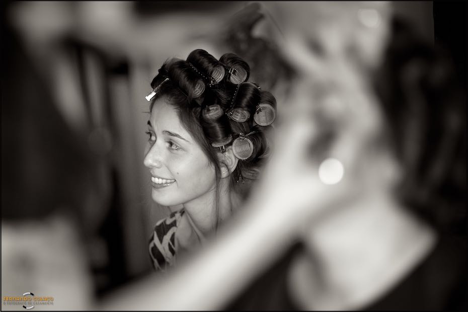 Vista no meio de elementos muito desfocados, a noiva, com rolos no cabelo, sorri para alguém num momento captado pelo fotógrafo de casamento em Sintra.