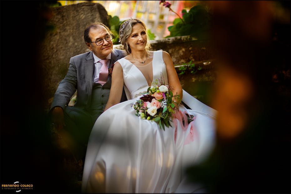 Sentados numa escadaria muito antiga e vistos por entre os troncos de árvores, desfocadas, do jardim da Quinta da Serra em Sintra, os noivos posam para o fotógrafo de casamento.