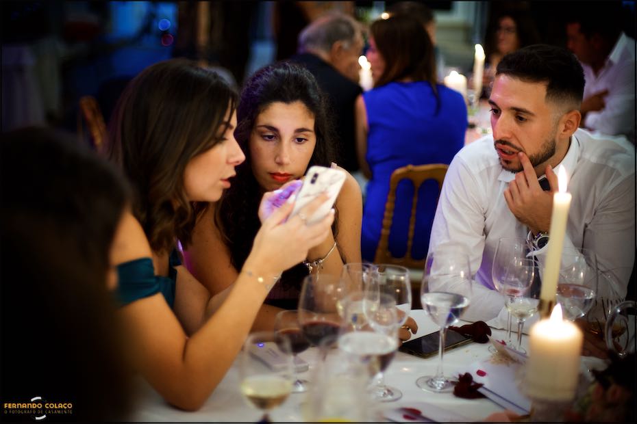 Numa das mesas do jantar da festa do casamento na Quinta do Pé da Serra em Sintra, duas amigas olham para algo num telemóvel, enquanto um amigo observa atentamente.