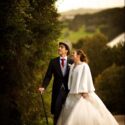In the session with the wedding photographer, the bride next to the groom, with a walking stick and tall hat, next to a large tree in the garden of Casa de Reguengos.