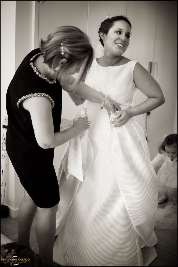 The bride's mother attaching a large bow to the waist of her wedding dress and a girl behind her adjusting her veil, seen by the wedding photographer in Seia, Portugal.